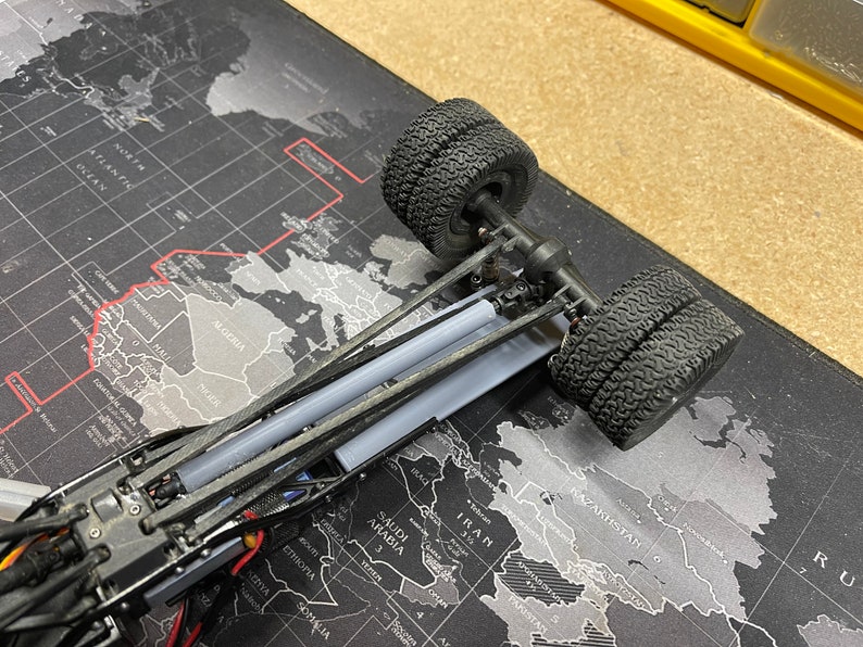 Op de afbeelding: Een close-up van een zwart speelgoedtruckchassis met twee zwarte banden. De truck staat op een zwart-grijze wereldkaart muismat.