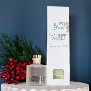 May include: A clear glass reed diffuser bottle with a gold cap and the text "Cranberry Woods" next to a white box with the same text. The box also has the "Blue Lava" logo and a small image of the product.