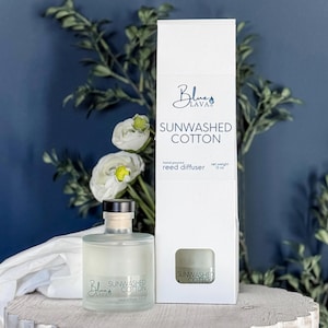 May include: A clear glass reed diffuser bottle and a white box with the text "SUNWASHED COTTON". The bottle has a wooden cap and contains a light-colored liquid. The box also has the text "Blue Lava" and "reed diffuser". The net weight is 12 oz.
