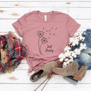 May include: A pink t-shirt with a black graphic of dandelions and the text "Just Dandy". The shirt is laid out on a white wooden surface with a plaid scarf, cotton bolls, and a pair of brown ankle boots.
