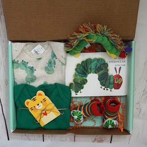 May include: A gift box filled with baby items, including a green and white onesie with caterpillar designs, a green long-sleeved onesie, a pair of colorful socks, and a book titled "The Very Hungry Caterpillar" by Eric Carle.