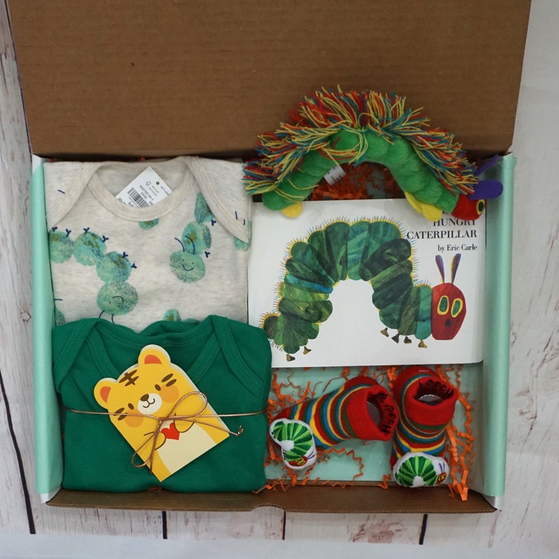 May include: A gift box filled with baby items, including a green and white onesie with caterpillar designs, a green long-sleeved onesie, a pair of colorful socks, and a book titled "The Very Hungry Caterpillar" by Eric Carle.