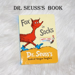 May include: A Dr. Seuss's Book of Tongue Tanglers board book featuring a red fox wearing blue socks. The book cover is yellow with the title "Fox in Socks" in blue and white text. The book is part of the Bright and Early Board Books series.
