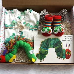 May include: A gift box filled with The Very Hungry Caterpillar themed baby items. The box is lined with white tissue paper and contains a white onesie with green caterpillars, a green plush caterpillar toy, a pair of red and green striped socks with caterpillar faces, and a copy of the book The Very Hungry Caterpillar by Eric Carle.