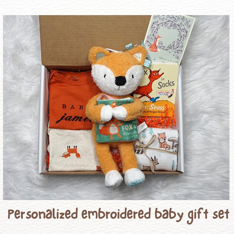 Puede incluir: Un set de regalo para beb&eacute; en una caja de cart&oacute;n que contiene un body naranja con el nombre James, una camiseta color crema con un gr&aacute;fico de zorro, un peluche de zorro naranja sosteniendo un libro, el libro Fox in Socks y una manta estampada.
