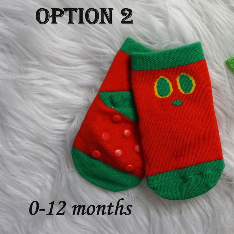 May include: Pair of red and green baby socks with non-slip grips on the bottom. One sock has a strawberry design and the other has a cartoon face with yellow eyes. The socks are labeled "0-12 months".