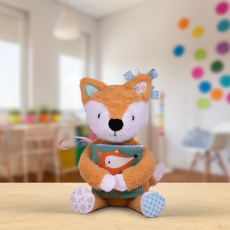 May include: A plush orange fox toy holding a book with a fox illustration on the cover. The book has the text "My Friends" on the bottom right corner. The fox toy has blue and white fabric accents and a pink ribbon.