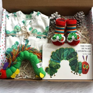 May include: A gift box filled with The Very Hungry Caterpillar themed baby items. The box is lined with white tissue paper and contains a green and red caterpillar plush toy, a white onesie with green caterpillars printed on it, a pair of red and green striped socks with caterpillar faces, and a copy of the book The Very Hungry Caterpillar by Eric Carle.