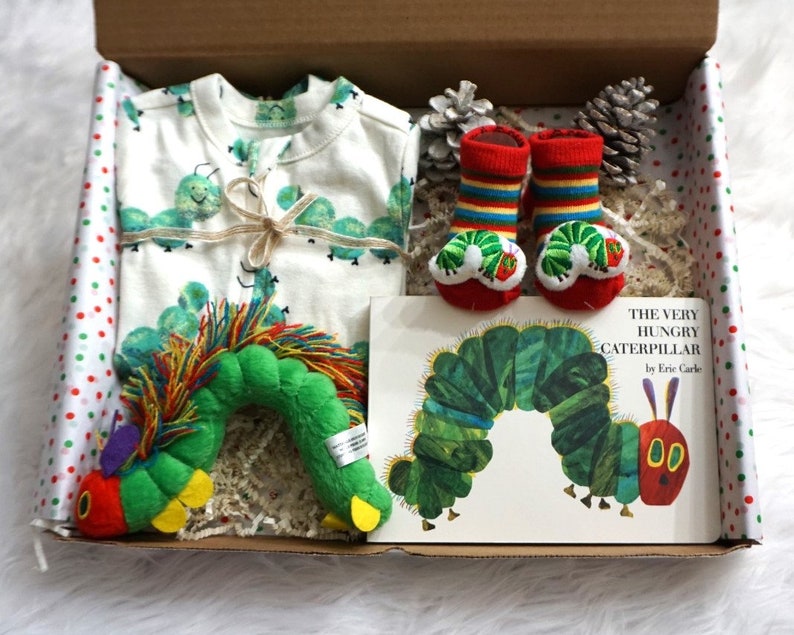 May include: A gift box filled with The Very Hungry Caterpillar themed baby items. The box is lined with white tissue paper and contains a green and red caterpillar plush toy, a white onesie with green caterpillars printed on it, a pair of red and green striped socks with caterpillar faces, and a copy of the book The Very Hungry Caterpillar by Eric Carle.