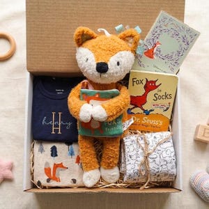 Puede incluir: Una caja de regalo que contiene un peluche de zorro naranja y blanco sosteniendo un libro peque&ntilde;o, un body azul marino con el nombre Henry bordado en oro, el libro Fox in Socks de Dr. Seuss, una manta con estampado de zorros y una tela blanca.
