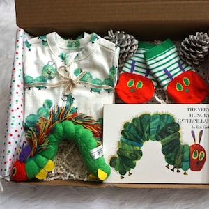 May include: A gift box filled with baby items themed around the book "The Very Hungry Caterpillar" by Eric Carle. The box includes a white onesie with green caterpillars, a green and white striped pair of socks with red caterpillar faces, a green caterpillar plush toy, and the book itself.