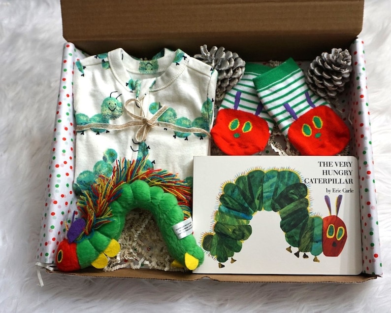 May include: A gift box filled with baby items themed around the book "The Very Hungry Caterpillar" by Eric Carle. The box includes a white onesie with green caterpillars, a green and white striped pair of socks with red caterpillar faces, a green caterpillar plush toy, and the book itself.