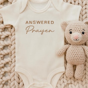 Könnte beinhalten: Cremefarbener Baby-Body mit den Worten "ANSWERED Prayer" in brauner Schrift gestickt. Ein gehäkelter Teddybär in passender cremefarbener Farbe sitzt neben dem Body. Die Artikel befinden sich auf einer strukturierten, cremefarbenen Decke.