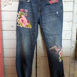 Puede incluir: Vaqueros azules de denim con parches florales bordados y detalles desgastados. Los vaqueros tienen un dobladillo vuelto y están decorados con flores de tela rosa y verde.