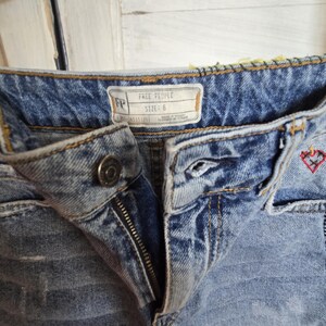 Peut inclure: Gros plan sur un short en jean bleu clair avec une fermeture &eacute;clair et un bouton. Le short pr&eacute;sente des broderies d&eacute;coratives et une &eacute;tiquette indiquant "FREE PEOPLE TAILLE: 6". Le jean a un aspect us&eacute;.