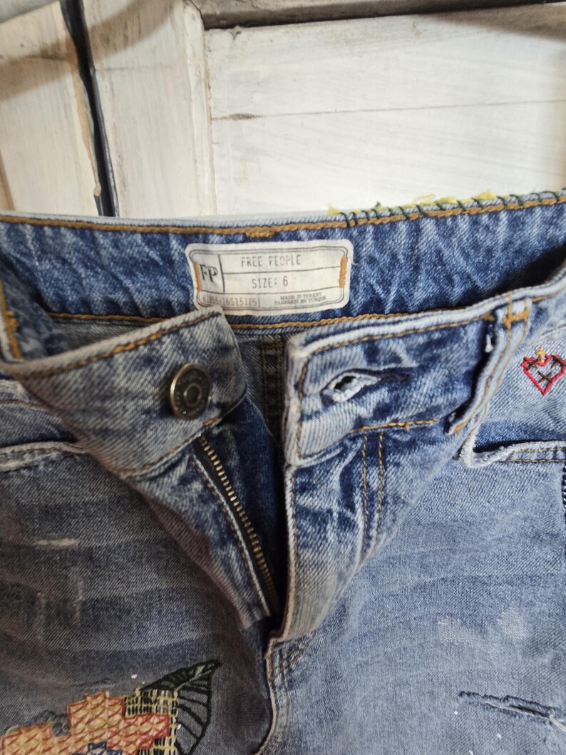 Peut inclure: Gros plan sur un short en jean bleu clair avec une fermeture &eacute;clair et un bouton. Le short pr&eacute;sente des broderies d&eacute;coratives et une &eacute;tiquette indiquant "FREE PEOPLE TAILLE: 6". Le jean a un aspect us&eacute;.
