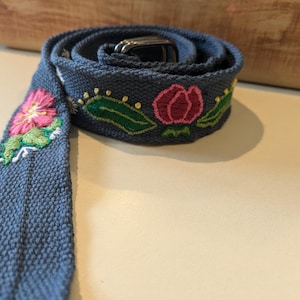 May include: A dark gray fabric belt with embroidered flowers in pink, green, and yellow. The belt has a metal buckle.