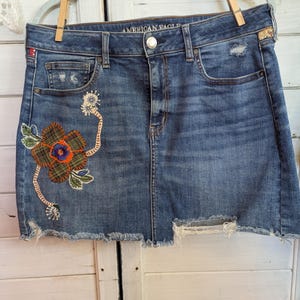 Può includere: Una gonna di jeans blu con orlo sfilacciato e ricami floreali. La gonna presenta dettagli usurati e la scritta "AMERICAN EAGLE" sulla cintura. Il ricamo floreale presenta un motivo a quadri con accenti arancioni e verdi.