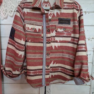 May include: A button-down shirt with a Southwestern-inspired pattern in shades of red, cream, and brown. The shirt features two chest pockets, a small patch on the sleeve, and a label that reads "BUFFALO TRACE". The cuffs are rolled up, revealing a striped lining.
