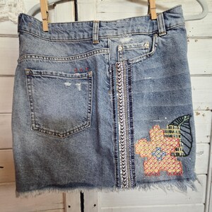 Peut inclure: Mini-jupe en jean bleu clair avec ourlet effiloch&eacute;, suspendue &agrave; un cintre gris. La jupe pr&eacute;sente un motif brod&eacute; de fleur et de feuille, et une bande verticale orn&eacute;e. Une poche arri&egrave;re est visible.