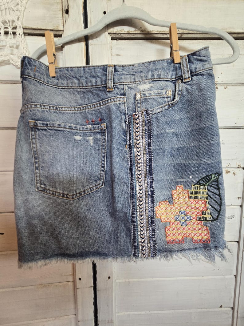 Peut inclure: Mini-jupe en jean bleu clair avec ourlet effiloch&eacute;, suspendue &agrave; un cintre gris. La jupe pr&eacute;sente un motif brod&eacute; de fleur et de feuille, et une bande verticale orn&eacute;e. Une poche arri&egrave;re est visible.