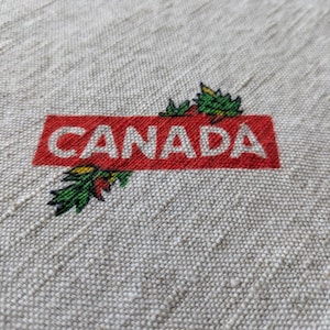 May include: Close-up of a beige fabric with a red rectangle that reads "CANADA" in white capital letters. Green and yellow leaf accents are on the left side of the rectangle. The fabric has a textured appearance.