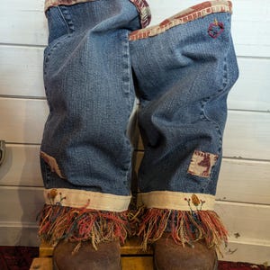Op de afbeelding: Een paar denim laarzen met bruine leren zolen en een franje rand. De laarzen zijn versierd met geborduurde bloemen en een patchwork ontwerp.