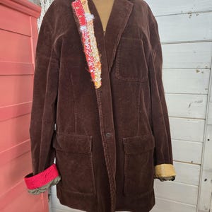 May include: A brown corduroy blazer with a decorative patch on the lapel. The blazer has two front pockets and the cuffs are rolled up to reveal a red and patterned lining. The blazer is displayed on a mannequin.