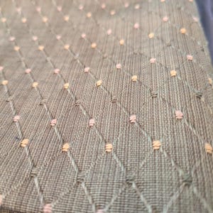 Pode incluir: Close-up de um tecido texturizado com um padrão de losangos. O tecido é de uma cor cinza-esverdeada suave, com pequenas costuras rosa e pêssego espaçadas uniformemente nas interseções do padrão de losangos. O tecido parece ser acolchoado.