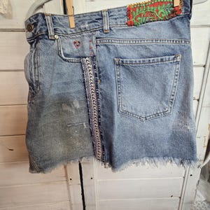 Peut inclure: Mini-jupe en jean bleu clair avec ourlet effiloch&eacute;, suspendue &agrave; un cintre. Comporte des coutures d&eacute;coratives, un d&eacute;tail de c&oelig;ur rouge et un patch avec broderie verte et rouge. La jupe a un aspect us&eacute;.