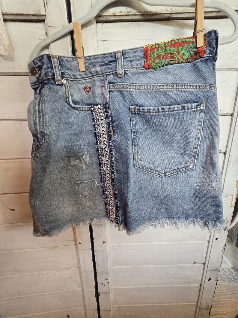 Peut inclure: Mini-jupe en jean bleu clair avec ourlet effiloch&eacute;, suspendue &agrave; un cintre. Comporte des coutures d&eacute;coratives, un d&eacute;tail de c&oelig;ur rouge et un patch avec broderie verte et rouge. La jupe a un aspect us&eacute;.