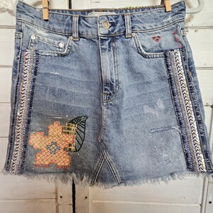 Peut inclure: Mini-jupe en jean bleu clair avec ourlet effiloch&eacute;, orn&eacute;e de broderies florales et de feuilles. La jupe pr&eacute;sente une garniture de perles d&eacute;coratives sur les c&ocirc;t&eacute;s et est suspendue &agrave; un cintre avec une pince &agrave; linge en bois.