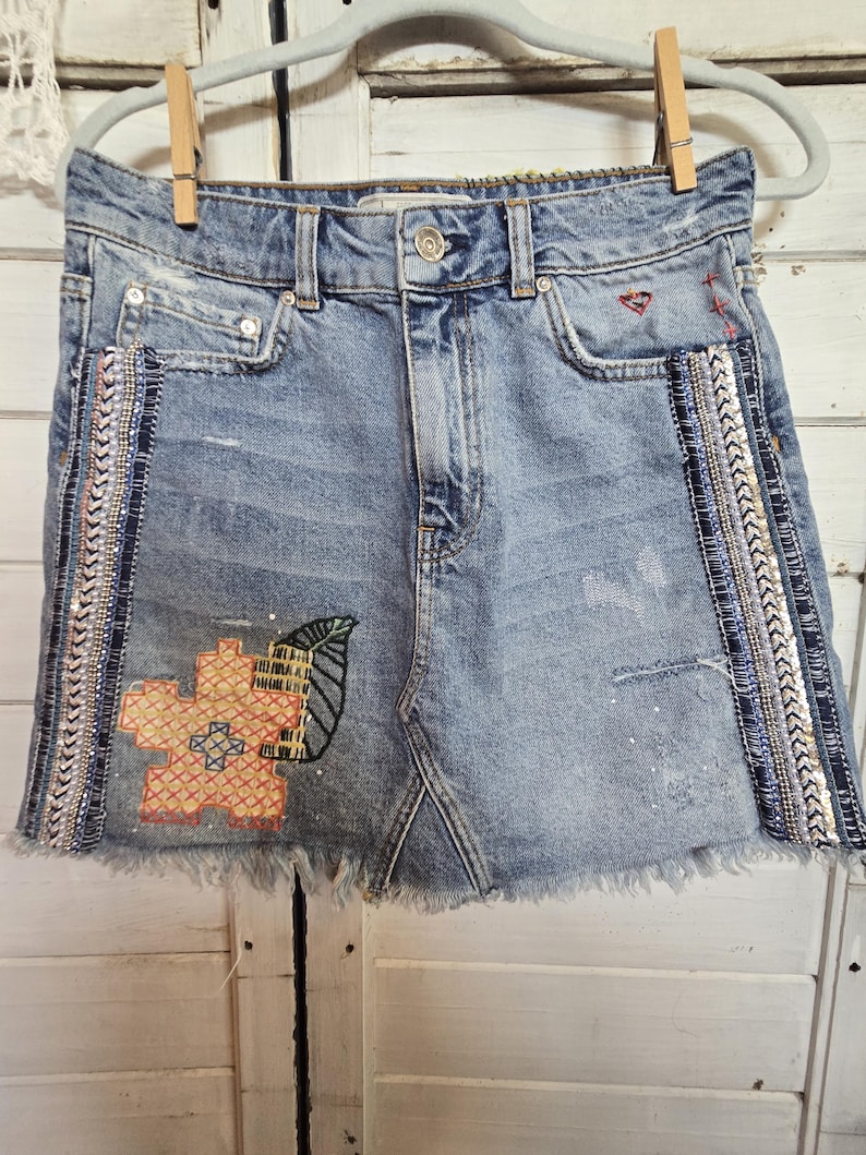Peut inclure: Mini-jupe en jean bleu clair avec ourlet effiloch&eacute;, orn&eacute;e de broderies florales et de feuilles. La jupe pr&eacute;sente une garniture de perles d&eacute;coratives sur les c&ocirc;t&eacute;s et est suspendue &agrave; un cintre avec une pince &agrave; linge en bois.