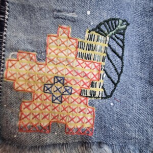 Peut inclure: Gros plan sur une veste en jean avec broderie au point de croix color&eacute;e. La broderie pr&eacute;sente un motif floral rouge, jaune et bleu, ainsi qu'une feuille verte et un motif de b&acirc;timent. La veste a un ourlet effiloch&eacute;.