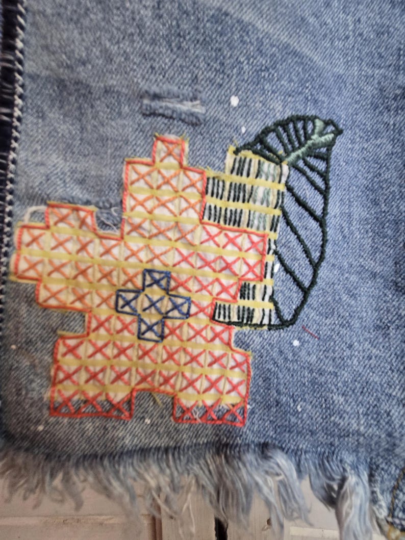 Peut inclure: Gros plan sur une veste en jean avec broderie au point de croix color&eacute;e. La broderie pr&eacute;sente un motif floral rouge, jaune et bleu, ainsi qu'une feuille verte et un motif de b&acirc;timent. La veste a un ourlet effiloch&eacute;.