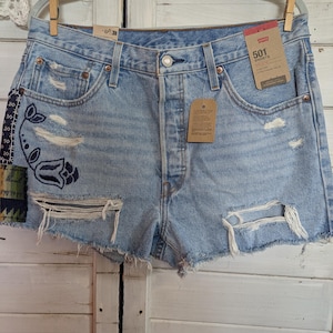 Könnte beinhalten: Hellblaue Jeansshorts mit Used-Details und einem dekorativen Aufnäher auf der linken Seite. Die Shorts haben einen Knopfverschluss und Taschen. Ein Etikett lautet "501 Shorts".