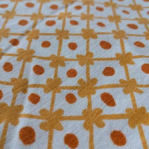 May include: Close-up of a white fabric with a repeating pattern of orange flower-like shapes and orange polka dots. The pattern creates a grid-like design, with the flowers at the intersections and dots filling the spaces.