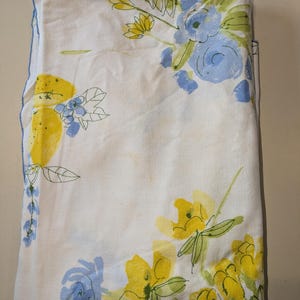 May include: A white tablecloth with a floral pattern in shades of yellow and blue. The tablecloth is folded in half and has a slightly distressed look.