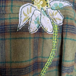 May include: A close-up of a plaid fabric with a large, handmade flower applique. The flower has white petals with green and blue accents, a yellow-green center, and a neon green stem. The plaid is in shades of green, brown, and blue.