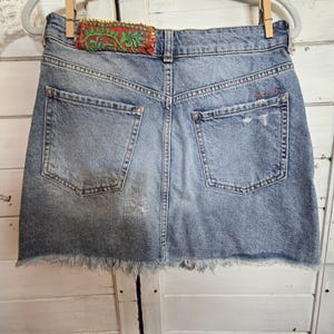 Peut inclure: Mini-jupe en jean bleu clair avec ourlet effiloch&eacute;, d&eacute;tails us&eacute;s et poches arri&egrave;re. La ceinture pr&eacute;sente un patch d&eacute;coratif avec broderie rouge et verte. La jupe est suspendue &agrave; un cintre gris avec des pinces &agrave; linge en bois.