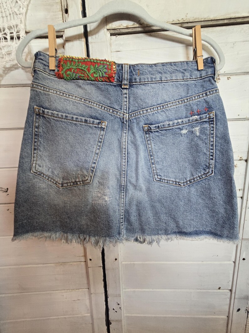 Peut inclure: Mini-jupe en jean bleu clair avec ourlet effiloch&eacute;, d&eacute;tails us&eacute;s et poches arri&egrave;re. La ceinture pr&eacute;sente un patch d&eacute;coratif avec broderie rouge et verte. La jupe est suspendue &agrave; un cintre gris avec des pinces &agrave; linge en bois.