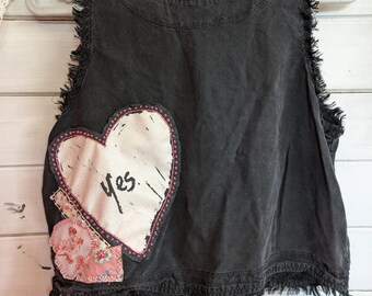 May include: A sleeveless black top with frayed edges and a round neckline. A large white heart with the word "yes" is appliqued on the front, along with a smaller patch of pink floral fabric. The top is hanging on a white hanger.