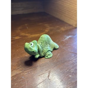 May include: A small, light green turtle figurine with a textured shell featuring carved geometric patterns. The turtle has a smooth head with visible white and black eyes, a slightly raised neck, and four distinct feet resting on the surface.