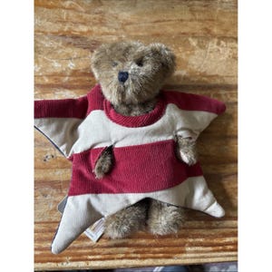 May include: A brown plush teddy bear with a dark nose, wearing a star-shaped garment made of red corduroy and beige fabric. The star-shaped piece features horizontal stripes and has openings for the bear's head and small furry arms to emerge.