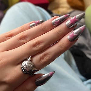 Könnte beinhalten: Silberne Chromnägel mit rosa Sterndesigns. Ein silberner Ring mit Stern- und Stacheldesign befindet sich am Ringfinger. Die Nägel sind lang und spitz.
