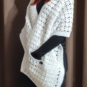 May include: A white crochet shawl with a V-neck design and two pockets. The shawl is made of a textured, open-weave pattern and features a button closure on one pocket. The garment is worn over a black top.