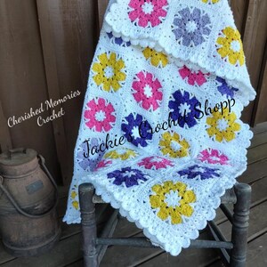 May include: White crochet baby blanket with a scalloped border and colorful flower motifs in pink, purple, yellow, and white. The blanket is draped over a wooden rocking chair. "Cherished Memories Crochet" and "Jackie's Crochet Shop" are written on the blanket.