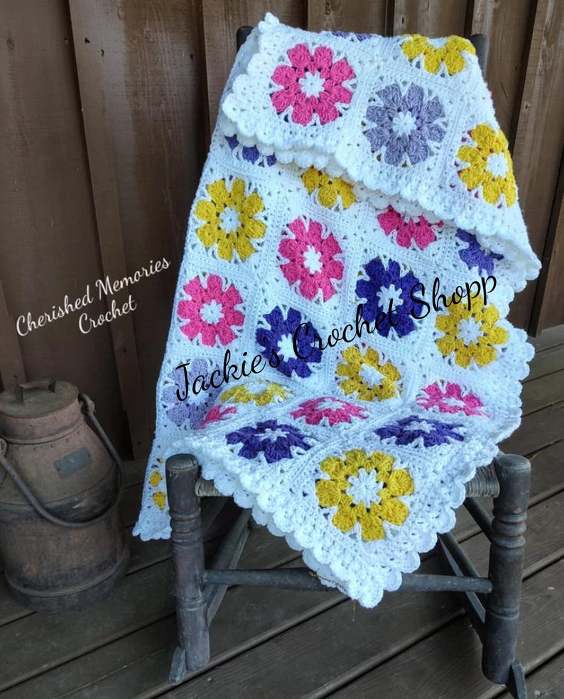 May include: White crochet baby blanket with a scalloped border and colorful flower motifs in pink, purple, yellow, and white. The blanket is draped over a wooden rocking chair. "Cherished Memories Crochet" and "Jackie's Crochet Shop" are written on the blanket.