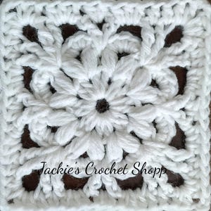 Puede incluir: Cuadrado de crochet blanco con un diseño de flor en el centro. El cuadrado está hecho con un punto de crochet sencillo y tiene un borde de puntos de crochet doble. El texto "Jackie's Crochet Shop" es visible en el centro inferior de la imagen.