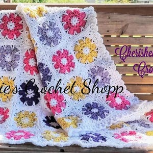 May include: A white crochet baby blanket with a scalloped edge. The blanket is made up of squares with different colored flowers in the center. The colors of the flowers are pink, yellow, purple, and white. The text "Jackies Crochet Shopp" and "Cherished Memories Crochet" is visible on the blanket.